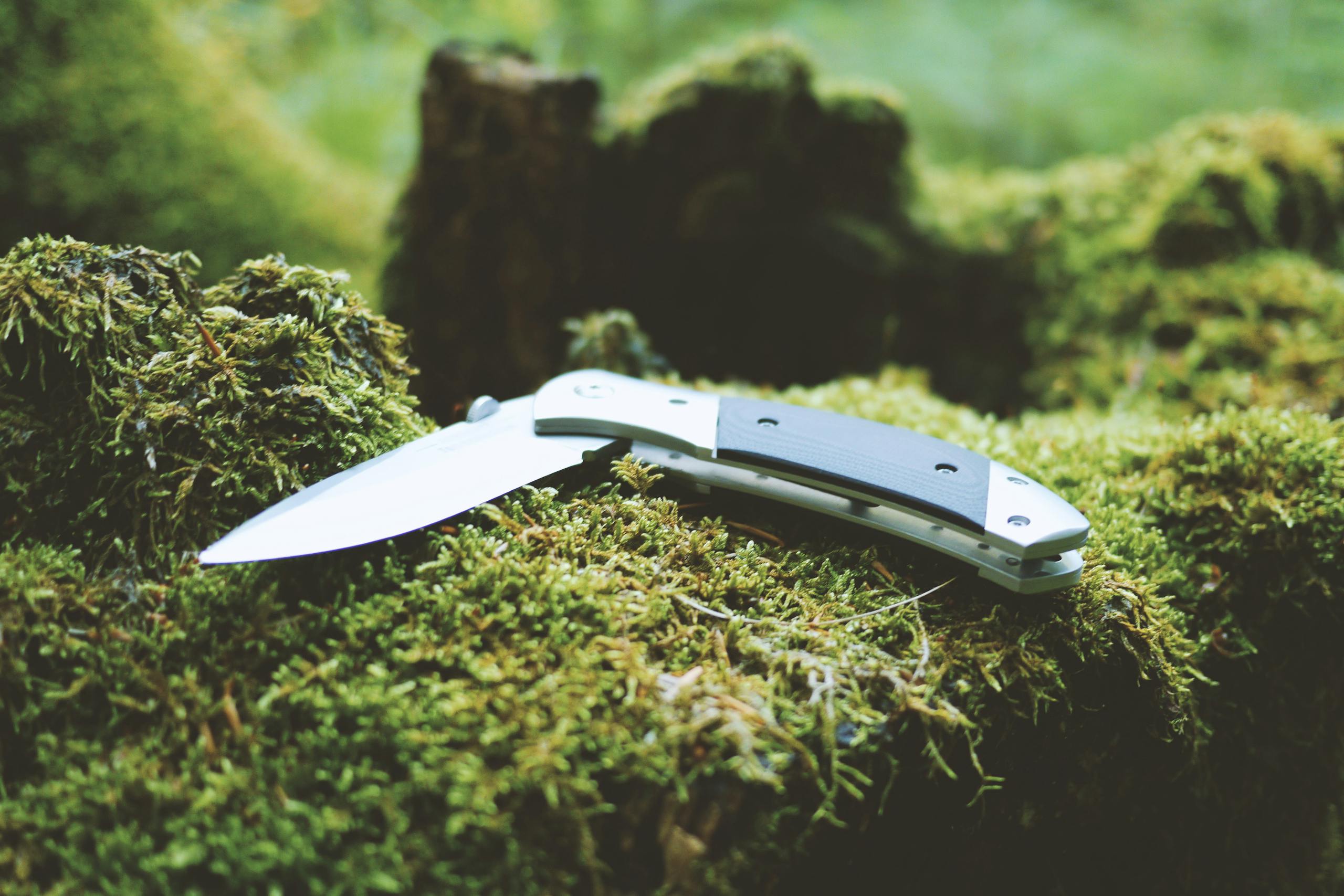 Folding knife resting on a lush moss-covered surface in a forest. Ideal for camping gear and outdoor enthusiasts.